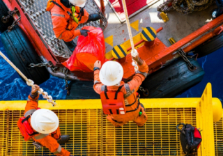 oil rig workers stand on a yellow metal platform, passing a bag over to other workers