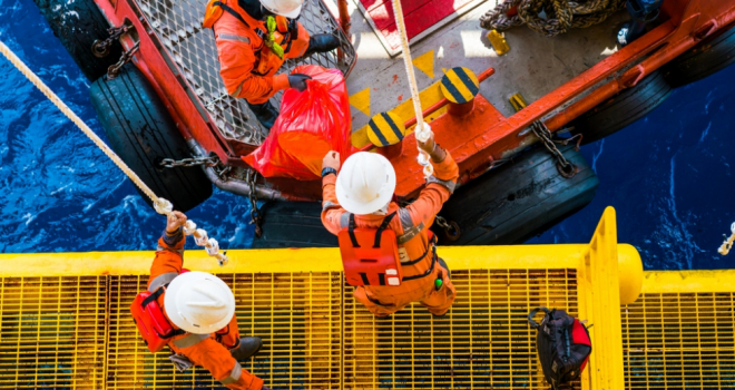 oil rig workers stand on a yellow metal platform, passing a bag over to other workers